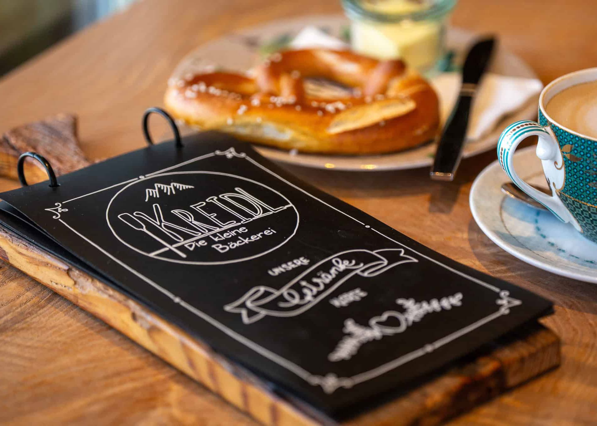 Gemütliche Impression im Kreidl Café: eine frische Brezel, Milchkaffee und das handgestaltete Mini-Kreidetafel-Menü auf einem Holzbrettchen.